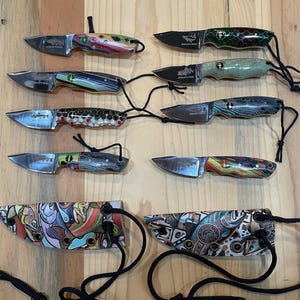 Op de afbeelding: Tien kleine messen met kleurrijke gepatroneerde handvatten en schedes. De messen hebben een zilveren blad en een zwart koord dat aan het handvat is bevestigd. De schedes zijn van leer en hebben een kleurrijk patroon.