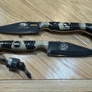 Puede incluir: Dos cuchillos plegables negros con mangos estampados y hojas negras se muestran sobre una superficie de madera. Un cuchillo tiene el texto "HORNWALT STEELHEAD" y el otro "ANGLERS EDGE" grabado en la hoja.