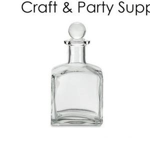 Puede incluir: Una botella de vidrio transparente con un tapón redondo. La botella está vacía y tiene forma cuadrada. La botella es para manualidades y artículos de fiesta.