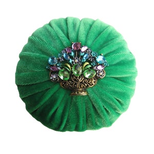 Puede incluir: Un cojín de alfileres redondo de terciopelo verde con un broche decorativo de piedras preciosas en el centro. El broche presenta una cesta dorada llena de brillantes de colores.
