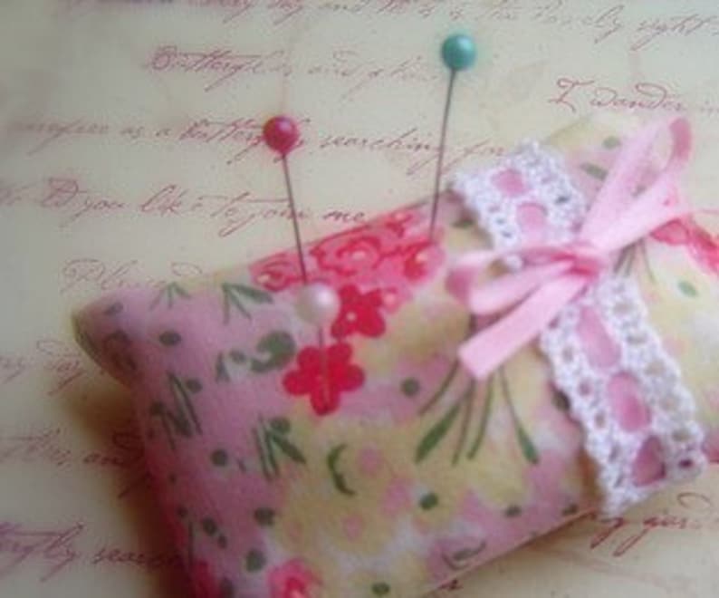 Puede incluir: Un coj&iacute;n para alfileres de tela floral rosa con una cinta rosa y un borde de encaje blanco. Tres alfileres est&aacute;n clavados en el coj&iacute;n, uno turquesa, uno rosa y uno blanco.