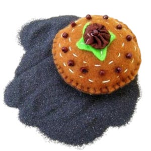 May include: A brown felt cake with a green and brown felt flower on top. The cake is surrounded by black sand.
