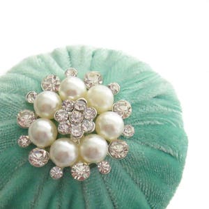 Puede incluir: Un cojín de alfileres de terciopelo azul turquesa con un gran adorno brillante de pedrería y perlas.