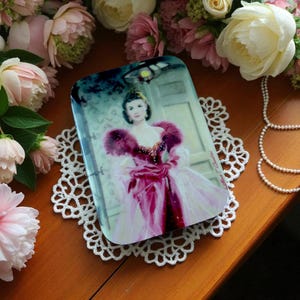 May include: Rectangular decorative tray featuring a portrait of a woman in a pink gown with a feathered shawl. The tray is set on a white lace doily, surrounded by pink and white flowers and a pearl necklace.