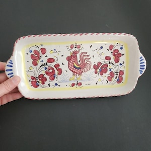 Puede incluir: Bandeja rectangular de cerámica con diseño de gallo y flores. La bandeja tiene una base blanca con borde amarillo y detalles rojos y azules. El gallo es rojo y amarillo con un patrón a cuadros.