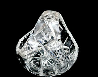 May include: A clear, cut-crystal basket with a handle. The basket features intricate geometric patterns and a starburst design on the base. The handle is arched and also cut with geometric shapes. The basket is set against a black background.