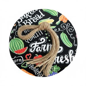 May include: A round black kitchen trivet with a colorful vegetable and fruit design. The trivet features a watermelon, corn, and other produce. The words "Baked Bread", "Farm Fresh", and "Quality" are written on the trivet. A twine bow is tied at the top.