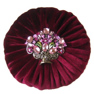 May include: A burgundy velvet round cushion with a decorative jeweled floral design in the center. The design features pink and clear rhinestones, green leaves, and a gold metal base.