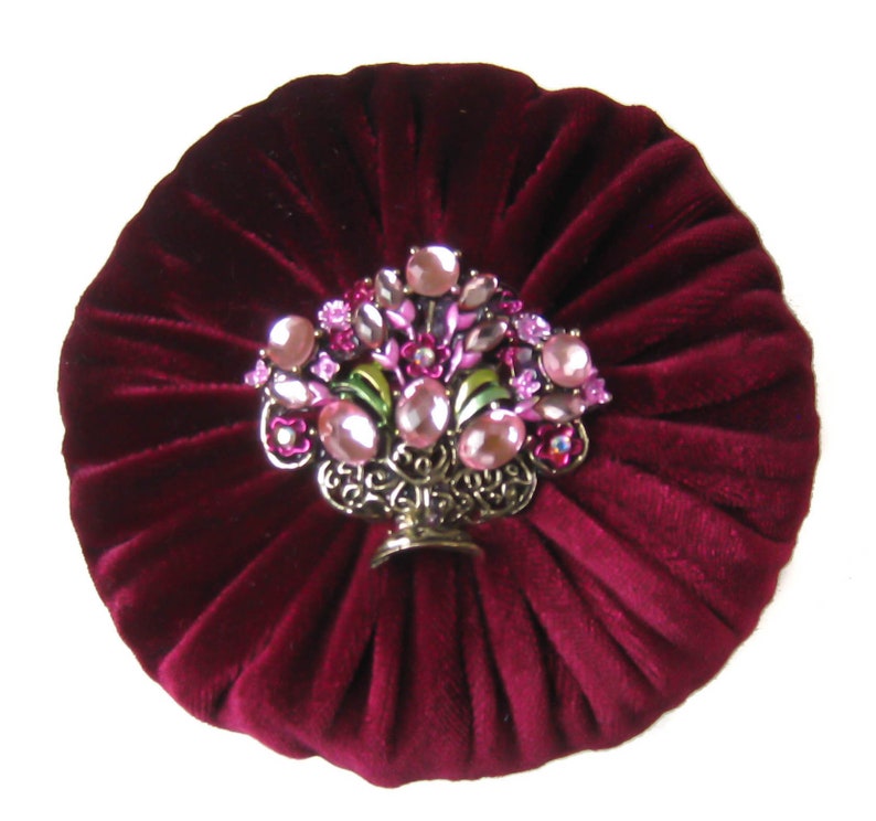 May include: A burgundy velvet round cushion with a decorative jeweled floral design in the center. The design features pink and clear rhinestones, green leaves, and a gold metal base.
