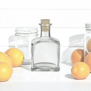 May include: A clear glass bottle with a cork stopper, a clear glass jar, and several oranges and lemons are arranged on a white surface. The bottle has a square shape and a cork stopper. The fruits are yellow and orange.