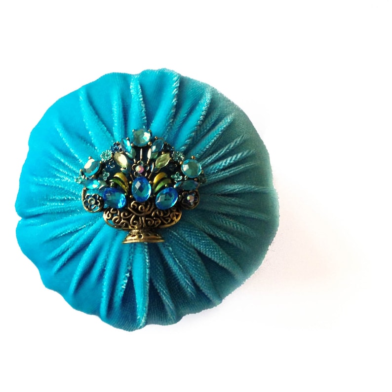 May include: A round, blue velvet cushion with a decorative jeweled brooch in the center. The brooch features a gold-toned basket overflowing with blue and green jewels.