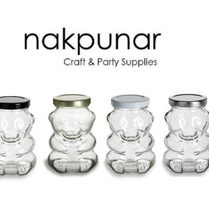 May include: Four clear glass bear-shaped jars with different colored lids. The lids are black, gold, silver, and silver. The jars are empty and have a smooth, clear glass surface. The jars are all the same size and shape. The jars are arranged in a row from left to right. The jars are on a white background. The text "nakpunar Craft & Party Supplies" is above the jars.