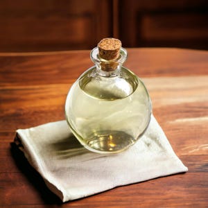 May include: A clear glass bottle with a cork stopper, filled with a light yellow liquid. The bottle is sitting on a white cloth napkin on a wooden surface.