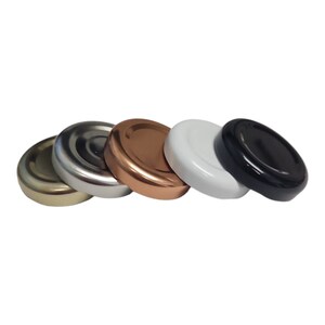 Puede incluir: Cinco tapones de botella de metal de diferentes colores. Los tapones están dispuestos en una fila de izquierda a derecha: dorado, plateado, cobre, blanco y negro.