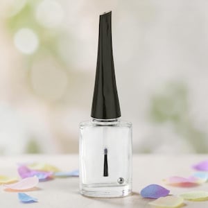 May include: A clear nail polish bottle with a black cap and brush applicator. The bottle is rectangular with a clear liquid inside. A small silver ball is visible at the bottom of the bottle. Colorful petals are scattered around the bottle.