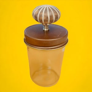 May include: A frosted glass jar with a brown lid and a decorative knob. The knob is a light brown and white colour with a raised design.