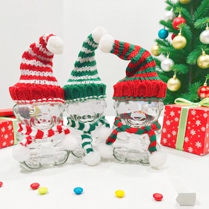 May include: Three clear glass bear-shaped jars, each adorned with a knitted Christmas hat and scarf. The hats are red and white striped, or green and white striped. The scarves are red and green striped. A Christmas tree and wrapped gift box are in the background.