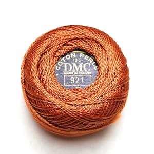 May include: A ball of orange DMC Coton Perle thread, 10g, with a blue label that reads "COTON PERLE DMC MADE IN FRANCE 921". The thread is tightly wound, showing the texture of the cotton. Ideal for embroidery and needlework.