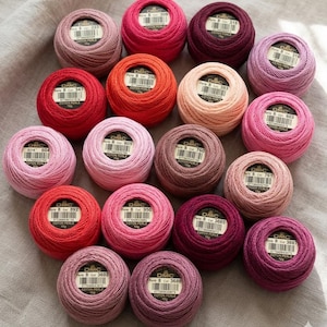 May include: A collection of DMC Perle Cotton thread balls in various shades of pink, red, and purple. Each ball has a label with the DMC logo and color information. The threads are arranged on a gray fabric surface.
