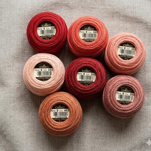 May include: Seven spools of DMC Perle cotton thread in various shades of red, pink, and orange. Each spool has a label with the DMC logo and color number. The threads are arranged on a gray fabric surface, ideal for embroidery and needlework projects.