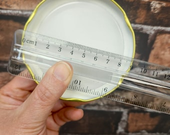 May include: A white metal lid with a gold rim, measured with a ruler. The ruler shows the lid is 7.5 cm in diameter.