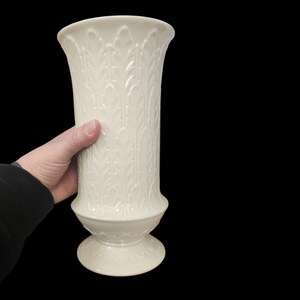 May include: A cream-colored, tall vase with a flared top and a pedestal base. The vase features a repeating leaf pattern and is held by a hand. The vase is approximately 10 inches tall.