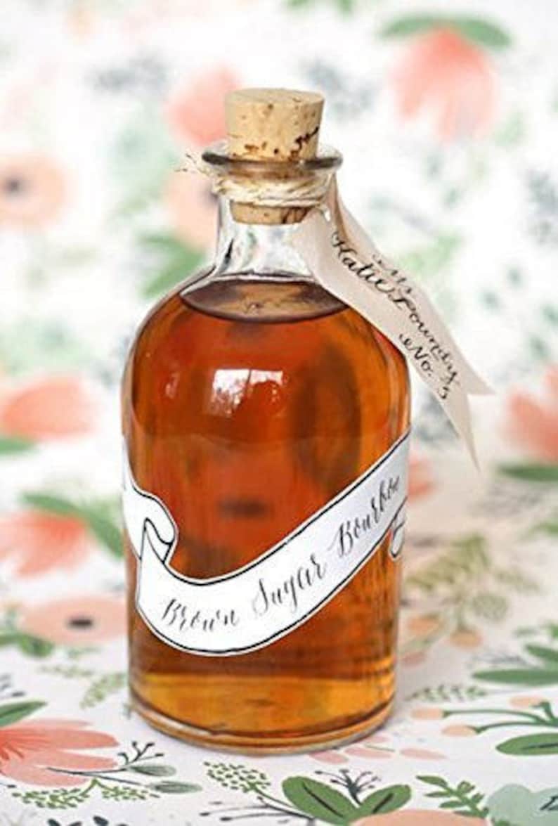 Puede incluir: Una botella de vidrio transparente llena de un l&iacute;quido marr&oacute;n, sellada con un tap&oacute;n de corcho. Una etiqueta blanca con las palabras "Brown Sugar Bourbon" envuelve la botella. Una etiqueta con "Katie Foundry No. 3" est&aacute; atada a la botella.