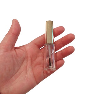 Puede incluir: Una botella de vidrio transparente con una tapa dorada y un aplicador de cepillo negro. La botella está llena de un líquido transparente.