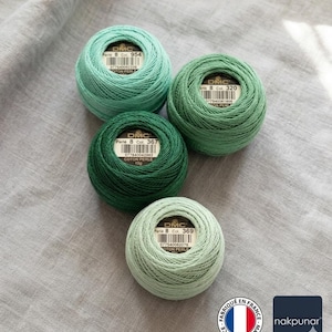 May include: Four balls of DMC Perle cotton thread in shades of green, from light to dark, are arranged on a gray fabric. Each ball has a label with the DMC logo and color information. The thread is tightly wound, showcasing the texture of the cotton.