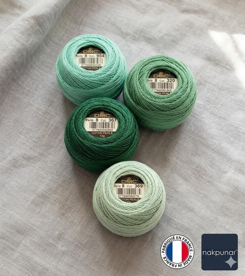 May include: Four balls of DMC Perle cotton thread in shades of green, from light to dark, are arranged on a gray fabric. Each ball has a label with the DMC logo and color information. The thread is tightly wound, showcasing the texture of the cotton.