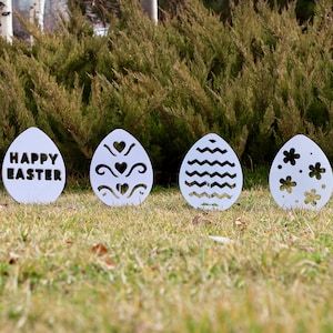 May include: Four white metal Easter egg decorations with different designs. The first egg says "Happy Easter". The second egg has a heart and swirl design. The third egg has a wavy line design. The fourth egg has a flower design.