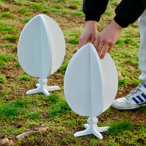 May include: Two white, freestanding, egg-shaped, decorative stands with a split design. The stands are on a grassy surface.