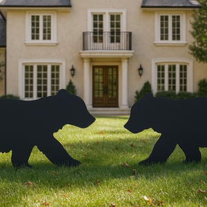 May include: Two black bear silhouette lawn decorations on a green lawn in front of a beige house. The bears are facing each other. The house has multiple windows and a dark roof.