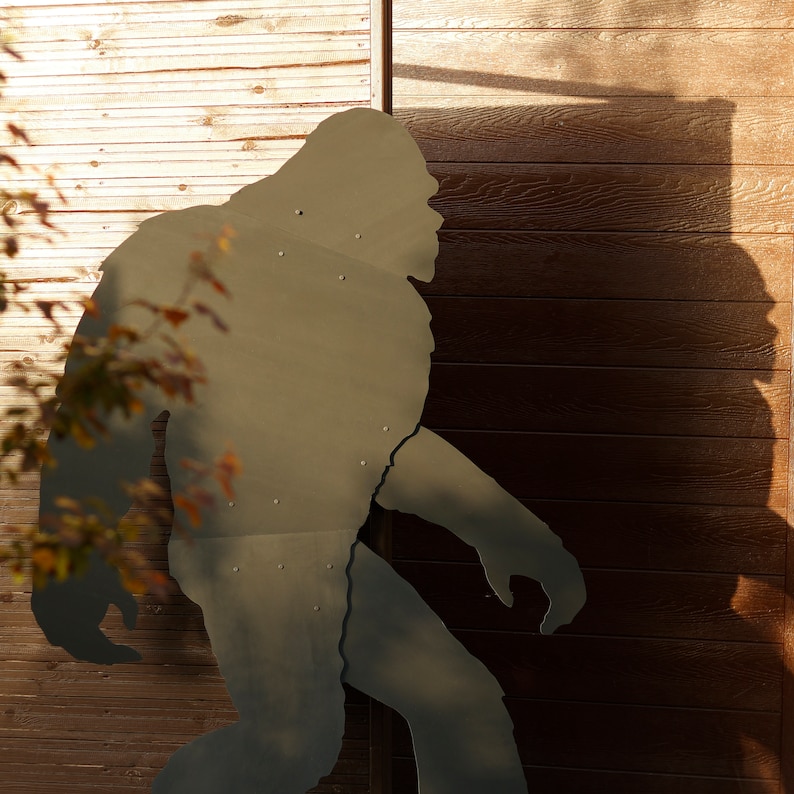May include: A silhouette of a large, hairy creature, possibly a Bigfoot, cut out of metal and mounted on a wooden wall.