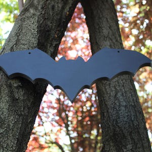 Op de afbeelding: Een zwart houten vleermuis silhouet hangend tussen twee boomstammen. De vleermuis is met spijkers aan de bomen bevestigd. De bomen bevinden zich in een bos met herfstbladeren op de achtergrond.