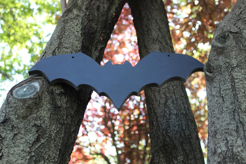 Op de afbeelding: Een zwart houten vleermuis silhouet hangend tussen twee boomstammen. De vleermuis is met spijkers aan de bomen bevestigd. De bomen bevinden zich in een bos met herfstbladeren op de achtergrond.