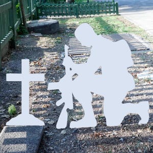 May include: A white silhouette of a soldier kneeling in prayer in front of a white cross. The soldier is holding a rifle in their right hand. The scene is set against a background of green grass and a wooden fence.
