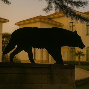Puede incluir: Silueta negra de un oso caminando sobre un muro de ladrillos, con una casa y un cielo crepuscular de fondo. El oso es el sujeto principal, con la casa y la vegetaci&oacute;n proporcionando contexto.
