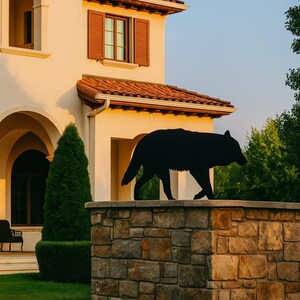 Puede incluir: Escultura de silueta de lobo de metal negro sobre un muro de piedra. La escultura est&aacute; frente a una casa beige con techo de tejas rojas y contraventanas marrones. Hierba verde y &aacute;rboles en el fondo.