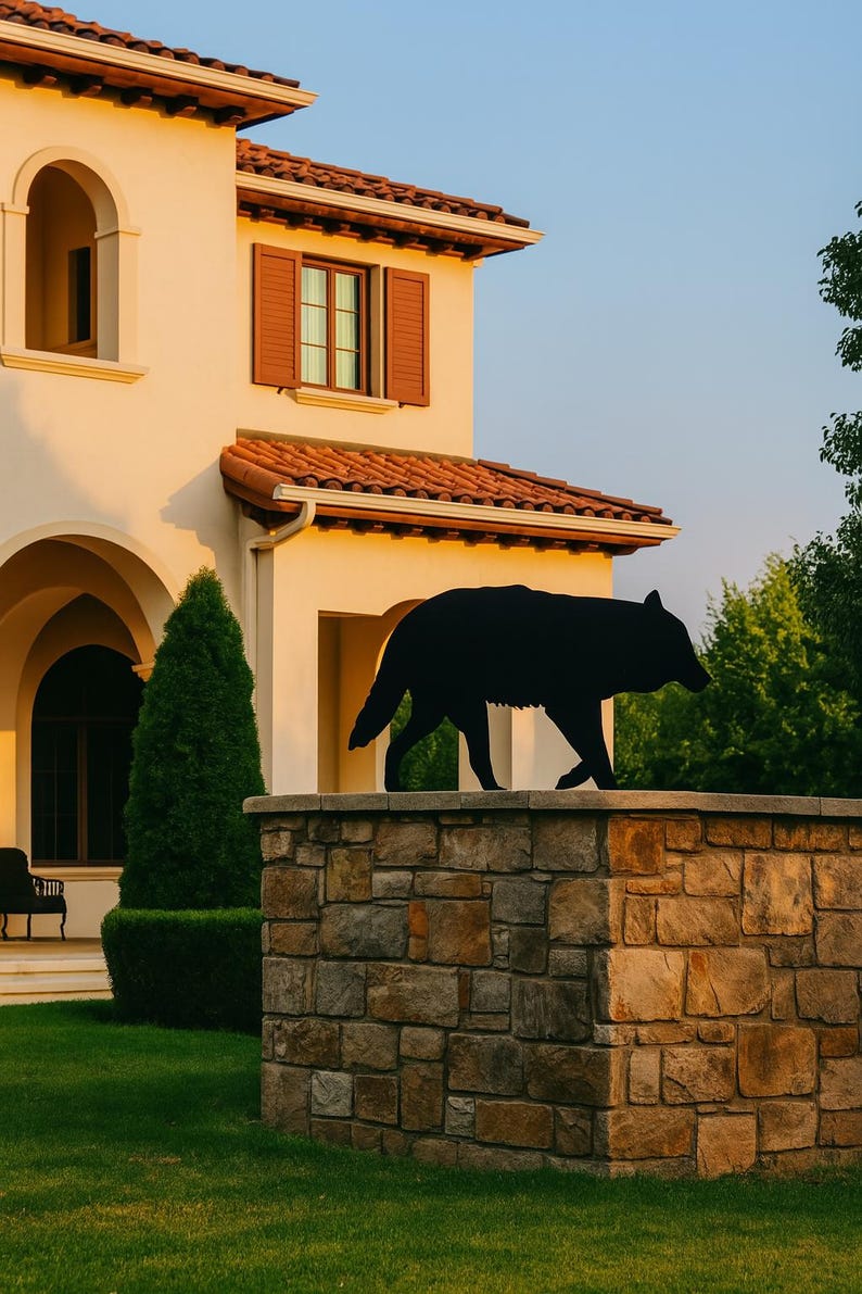 Puede incluir: Escultura de silueta de lobo de metal negro sobre un muro de piedra. La escultura est&aacute; frente a una casa beige con techo de tejas rojas y contraventanas marrones. Hierba verde y &aacute;rboles en el fondo.