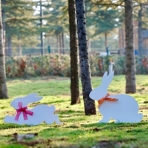 May include: Two white wooden bunny silhouettes with orange and pink ribbon bows on a grassy lawn in front of trees.