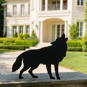 Puede incluir: Silueta met&aacute;lica negra de un lobo aullando, colocada sobre un muro de piedra. El fondo presenta una gran casa color crema con c&eacute;sped y &aacute;rboles bien cuidados, creando un contraste visual con la obra.