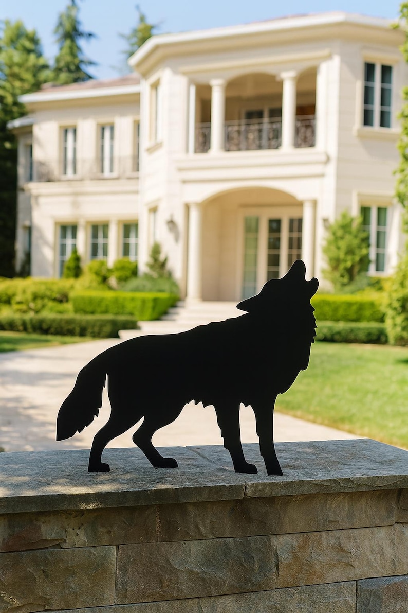 Puede incluir: Silueta met&aacute;lica negra de un lobo aullando, colocada sobre un muro de piedra. El fondo presenta una gran casa color crema con c&eacute;sped y &aacute;rboles bien cuidados, creando un contraste visual con la obra.