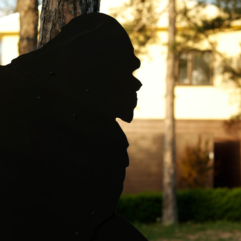 May include: A black silhouette of a person's head and shoulders, facing to the right, with a blurred background of a building and trees.