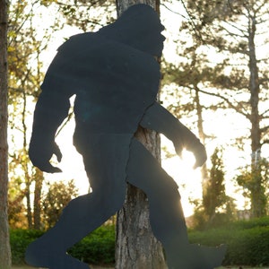 May include: A black silhouette of Bigfoot, a mythical creature, standing in front of a tree. The silhouette is cut out of metal and is attached to the tree with screws.