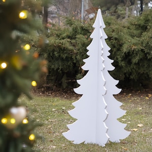 May include: A large, freestanding white Christmas tree decoration. The tree is made of cut-out panels and stands on a grassy surface. The tree is in front of a green bush. The image is taken outdoors.
