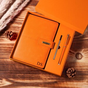 Könnte beinhalten: Ein orangefarbenes Leder-Notizbuch- und Stift-Set in einer Geschenkbox. Das Notizbuch hat einen Klappenverschluss mit silberner Schließe und die Initialen "DS" auf der Vorderseite. Ein silberner Stift und eine Stiftmine sind ebenfalls enthalten.