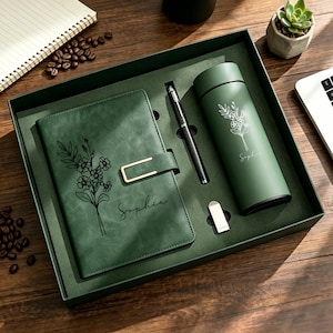 Può includere: Un set regalo verde scuro contenente un taccuino con chiusura dorata, una penna nera e un bicchiere termico abbinato. Sia il taccuino che il bicchiere presentano un'illustrazione floreale a linee bianche e il nome Sophia in corsivo.
