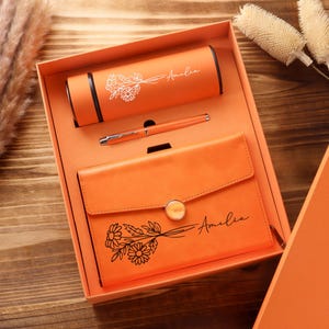 May include: An orange gift set in a box containing a cylindrical bottle, a ballpoint pen, and a notebook. The bottle and notebook feature a floral line art illustration and the name Amelia printed in a script font. The notebook has a button closure.