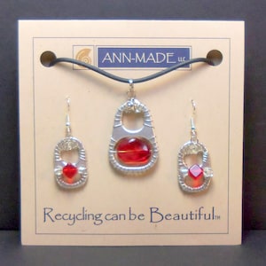 May include: A set of jewelry made from recycled materials. The set includes a necklace, a pair of earrings, and a pendant. The pendant and earrings are shaped like a can tab and feature a red glass bead. The necklace is black and has a silver clasp. The jewelry is displayed on a card with the text "ANN-MADE LLC" and "Recycling can be Beautiful TM".